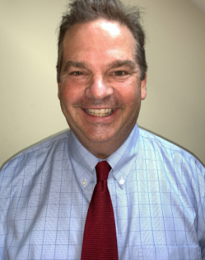 Chris Robertson smiles at the camera wearing a blue shirt and red tie