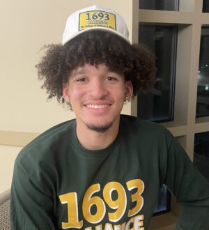 Ryan Jackson, Jr. smiles at the camera wearing a 1693 alliance shirt and hat.