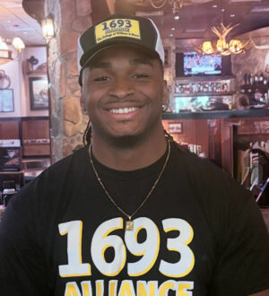 Jorden McDonald smiles while wearing a 1693 Alliance hat and t shirt